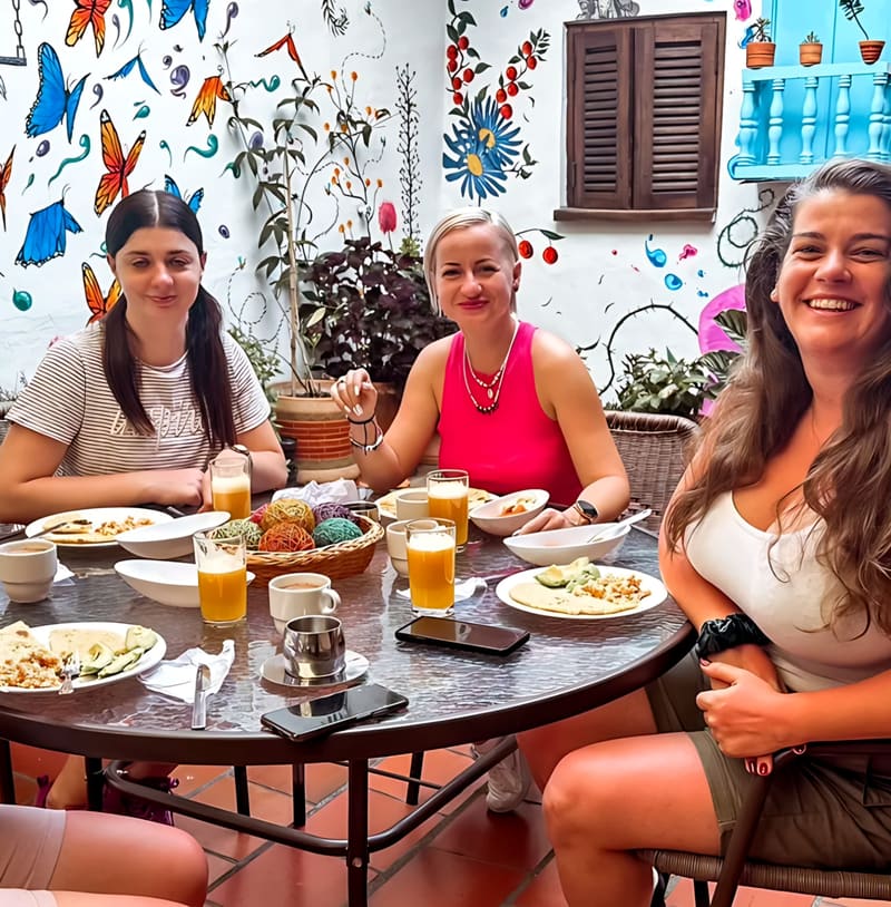 Tres huéspedes sonríen en el desayuno, con jugo de naranja, huevos y aguacate, en el patio de Real Dream frente al mural de mariposas y flores pintado a mano.