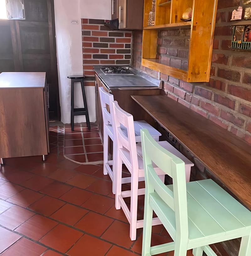 Barra del área de restaurante con sillas de madera pintadas de blanco, rosado y verde menta, muro de ladrillo y piso de terracota.