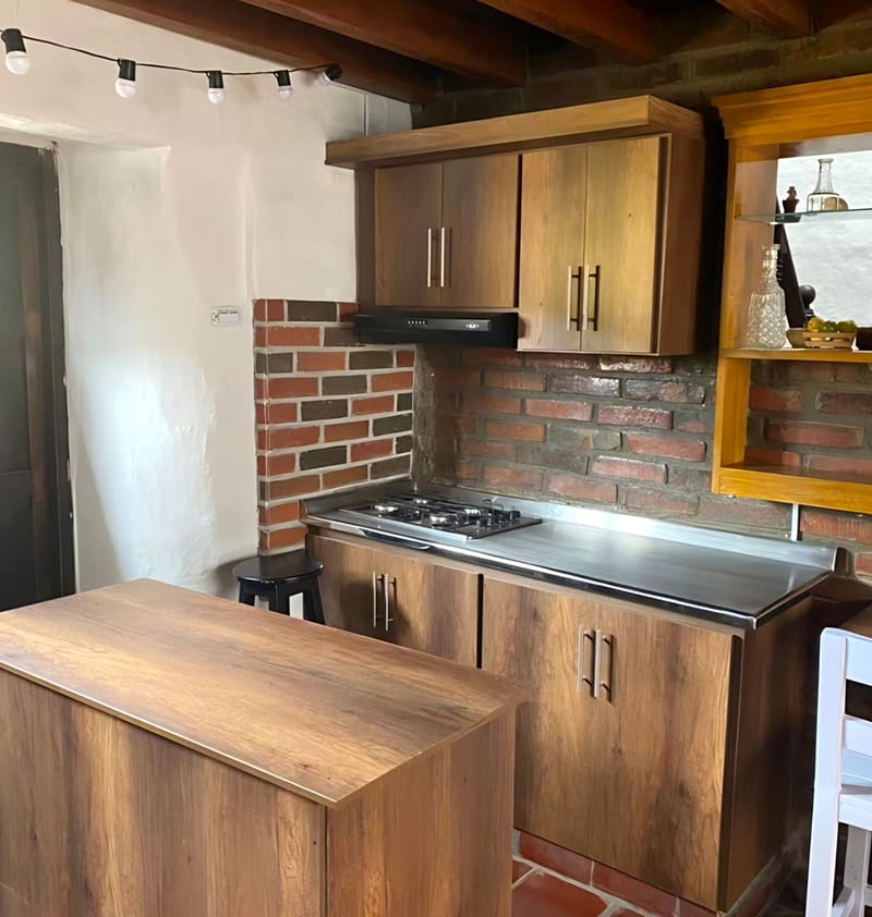 Cocina del área de restaurante con muebles de madera, estufa, campana y muro de ladrillo bajo una guirnalda de bombillos.