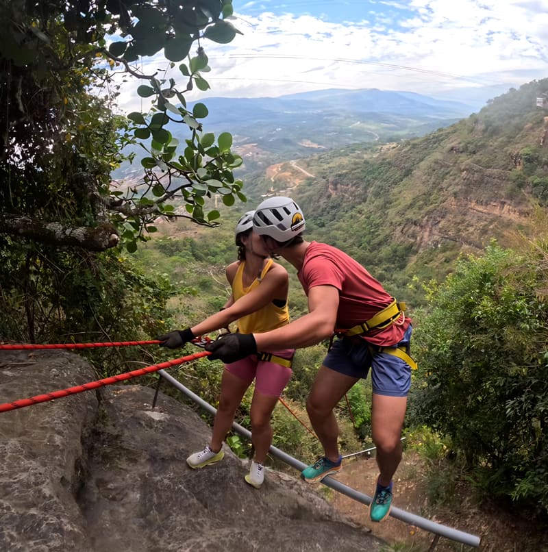 Dos personas con casco y arnés, sujetas a una cuerda roja al borde de una roca, comparten un beso antes de un rappel con el valle verde de Santander al fondo.