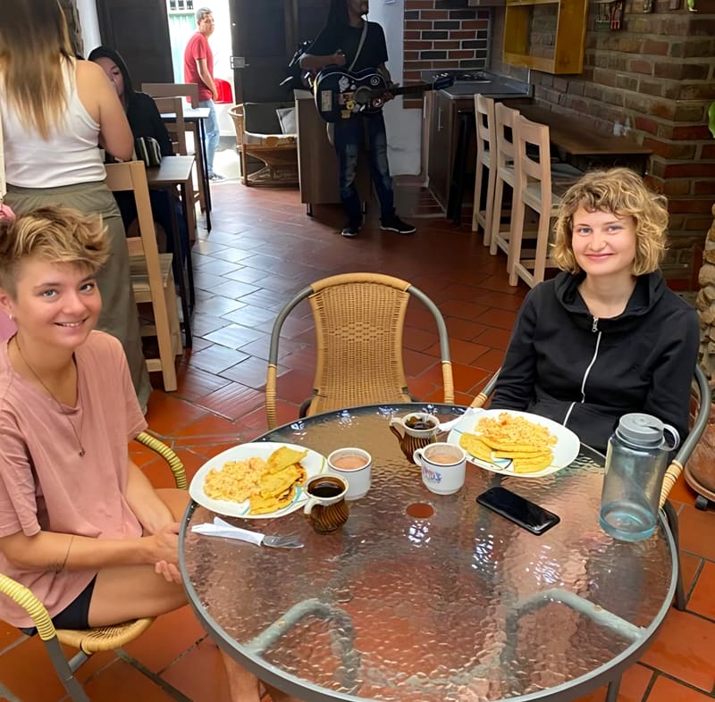 Dos huéspedes desayunan huevos revueltos en el área de restaurante mientras un músico toca la guitarra al fondo.