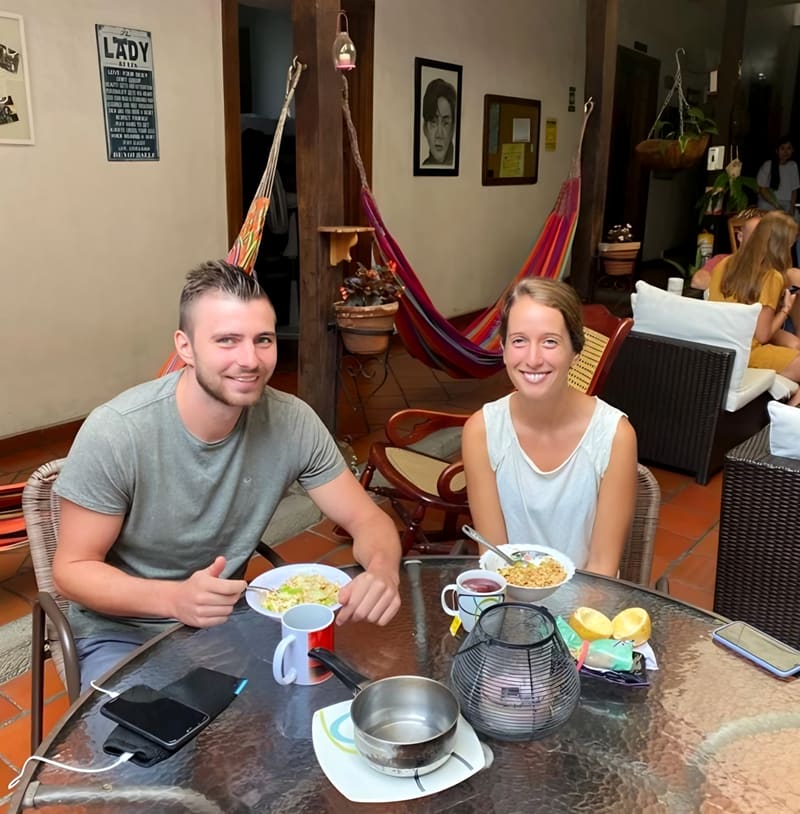Zona común con hamacas de colores colgadas de vigas de madera, sillas de mimbre y una pareja desayunando en una mesa de vidrio.
