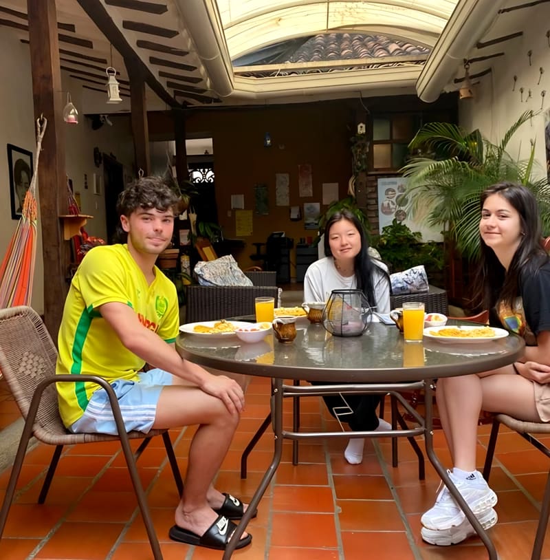 Patio cubierto con claraboya, vigas de madera, una hamaca y plantas; tres huéspedes desayunan con jugo de naranja.
