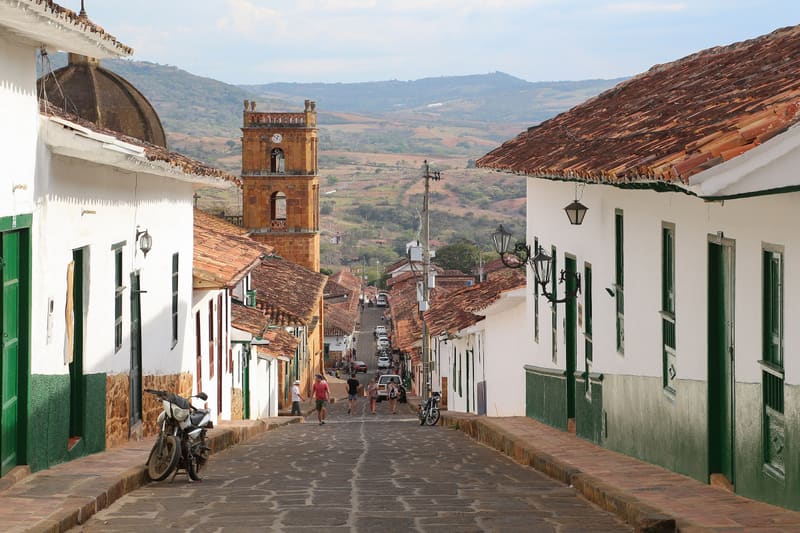 Calle empedrada de Barichara con casas blancas, puertas verdes y la torre de la catedral al fondo.
